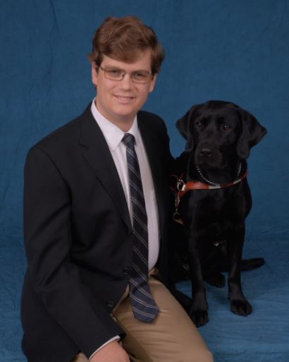Dr. Hasley and his guide dog, Shade. Photograph of Dr. Hasley wearing a tie and coat (blue). Black dog to his right. 