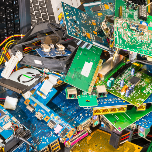 Circuit boards and discarded electronics. Green circuit boards on the right and blue ones on the bottom left. Keyboards are visible on the top right corner. 